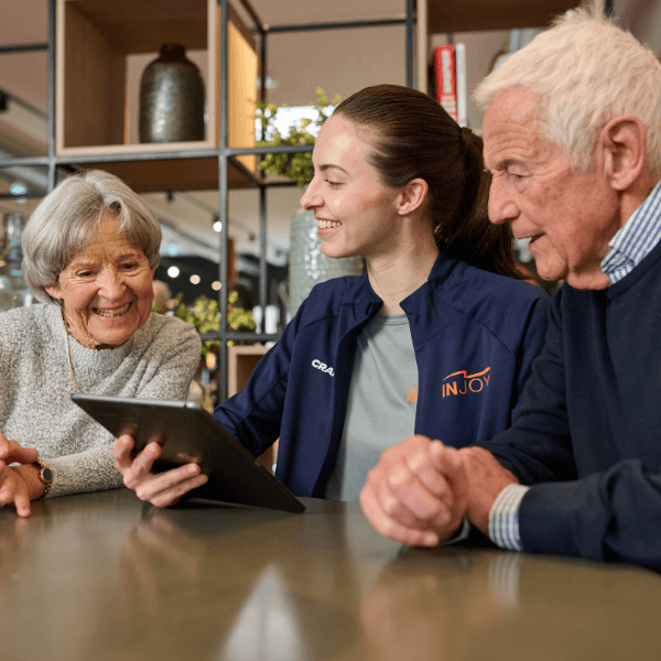 Freundliches älteres Ehepaar lässt sich von einer INJOY Trainerin mit iPad beraten.