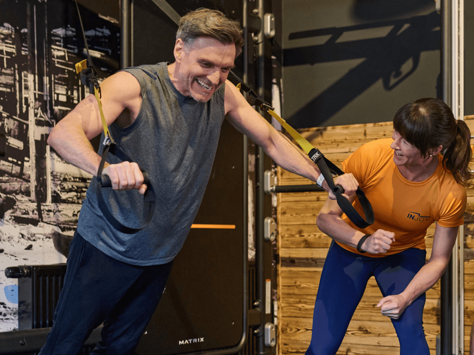 Ein männliches INJOY Mitglied trainiert an einem TRX im Fitnessstudio und wird dabei von einer Trainerin in orangem Shirt motiviert.