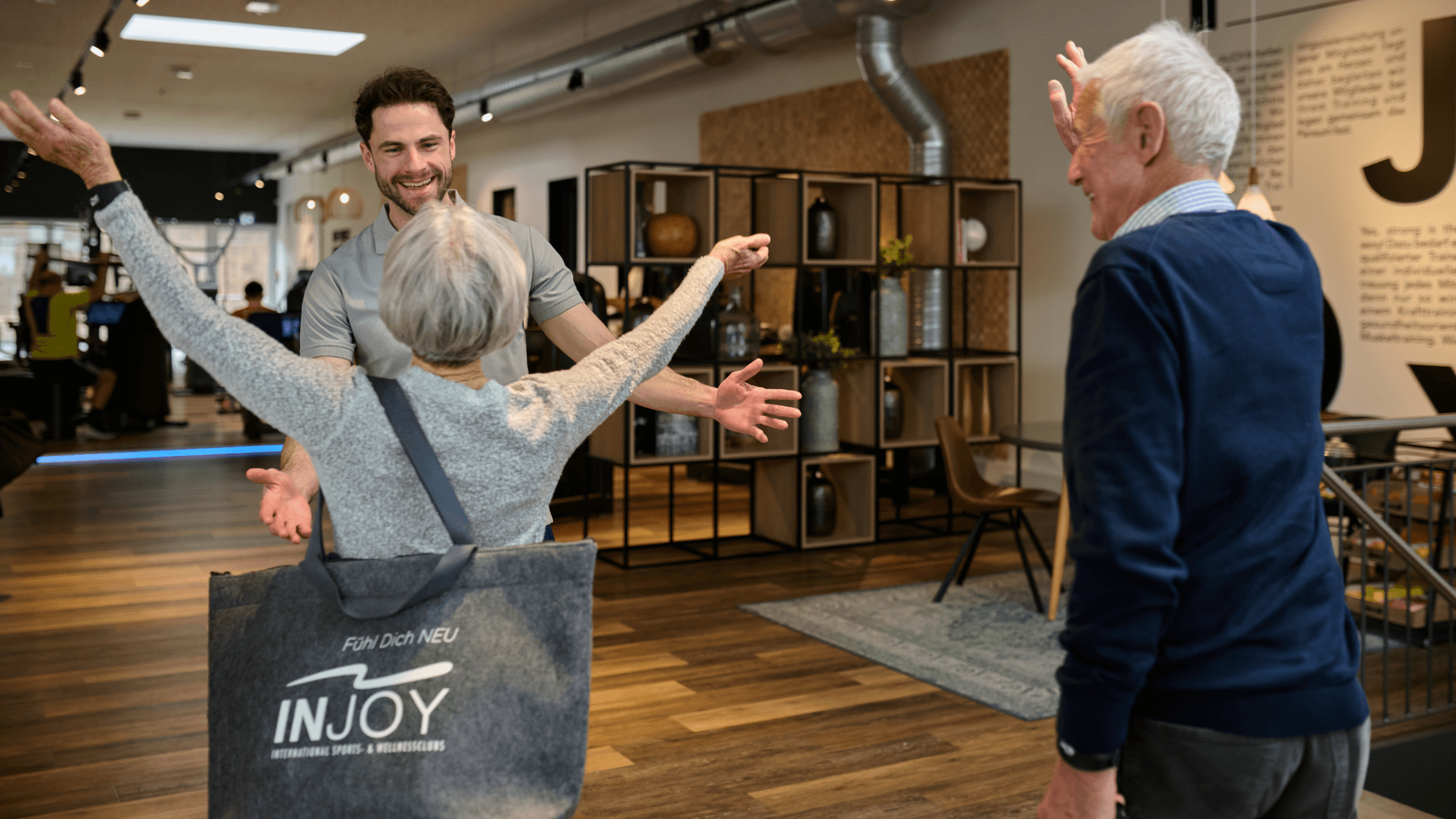 Zwei ältere Mitglieder werden von einem jungen INJOY Trainer herzlich im Fitnessstudio begrüßt.