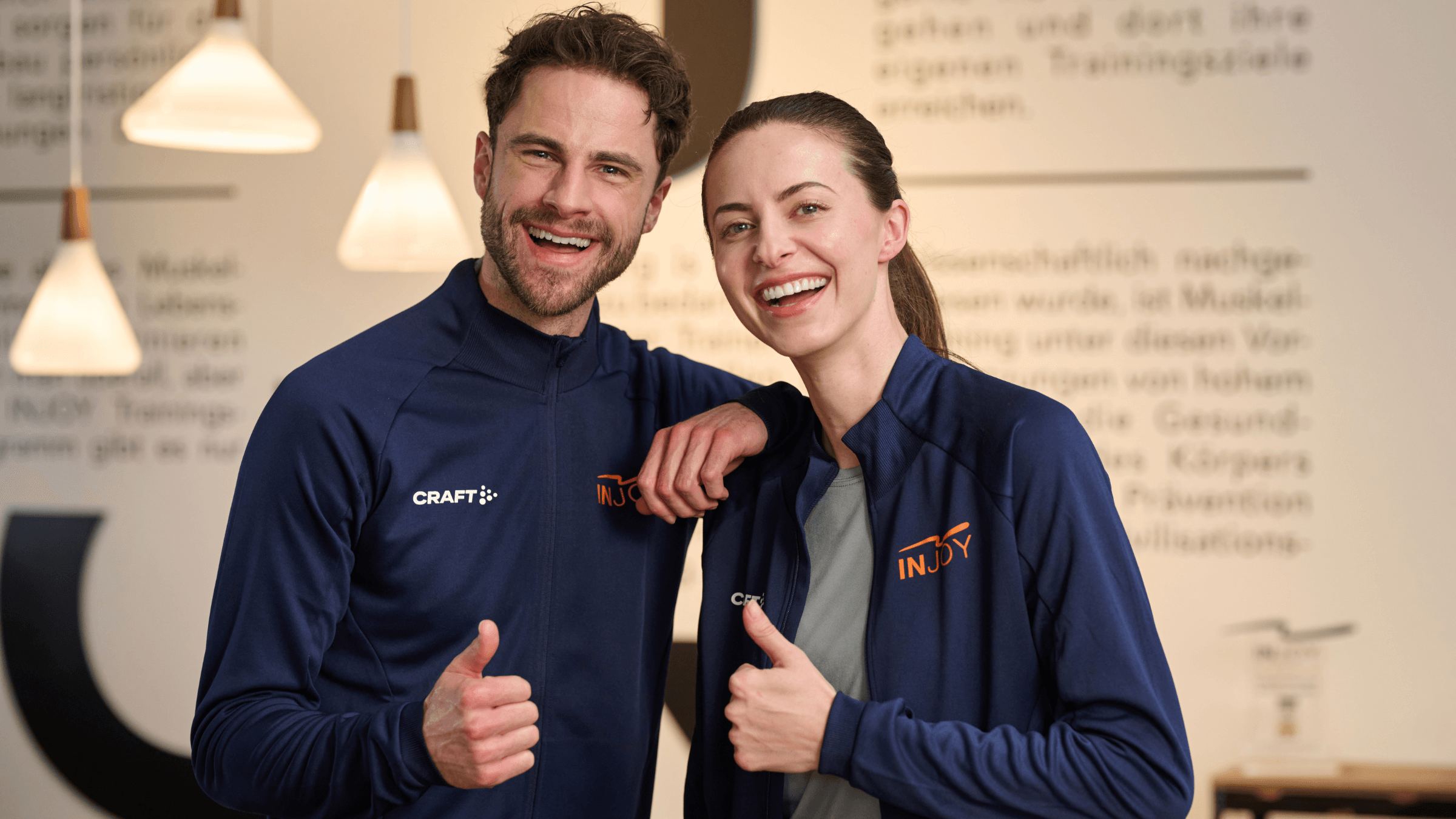 Ein männlicher INJOY Trainer und eine weibliche INJOY Trainerin stehen nebeneinander und geben einen Daumen nach oben.