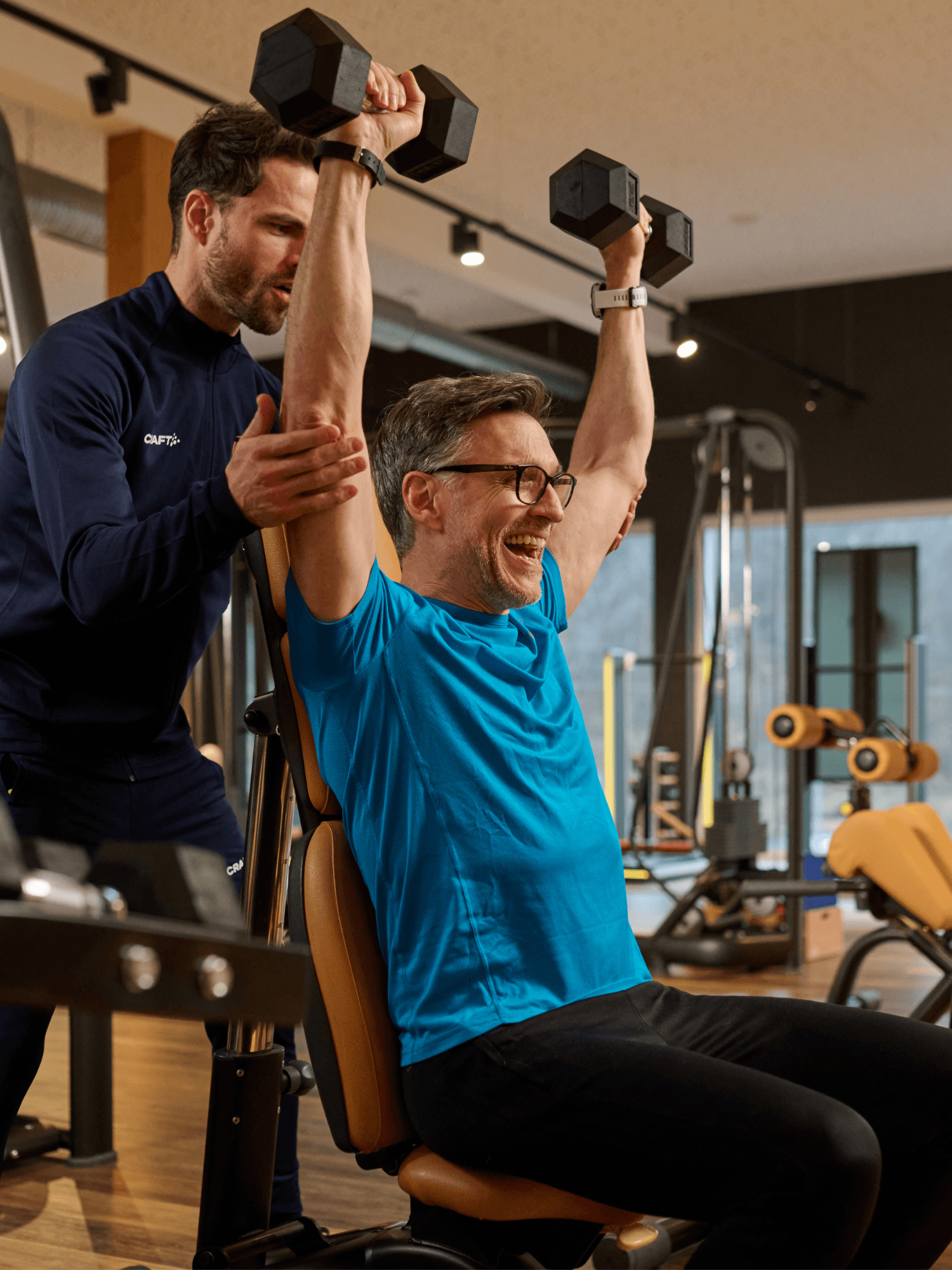 Ein INJOY Trainer steht hinter einem sitzenden Mitglied und hilft ihm bei der Ausführung einer Schulterübung.