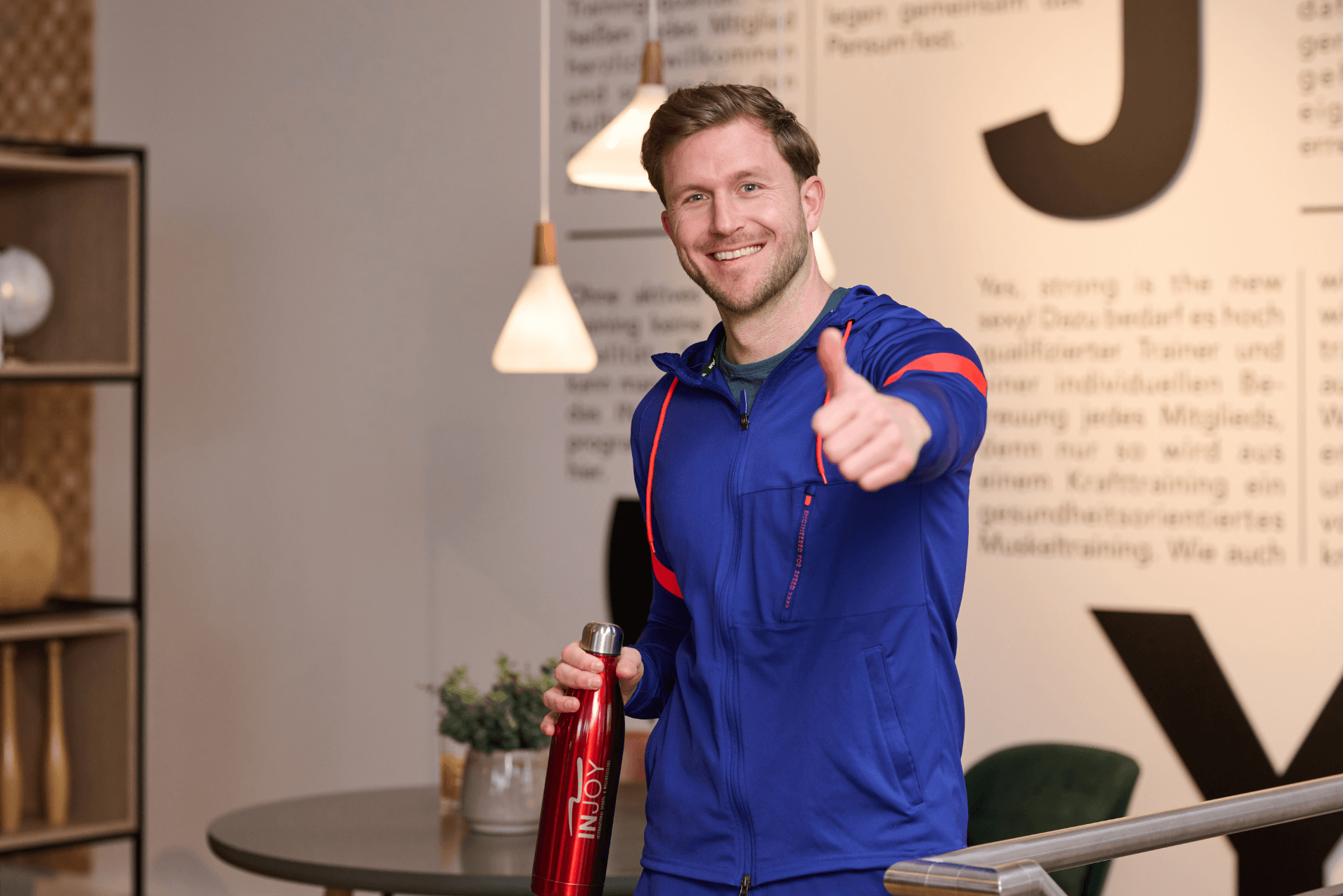 Der Mann in sportlicher Kleidung posiert mit Daumen hoch und einer Wasserflasche in der Hand, in einer modernen Fitnessumgebung.