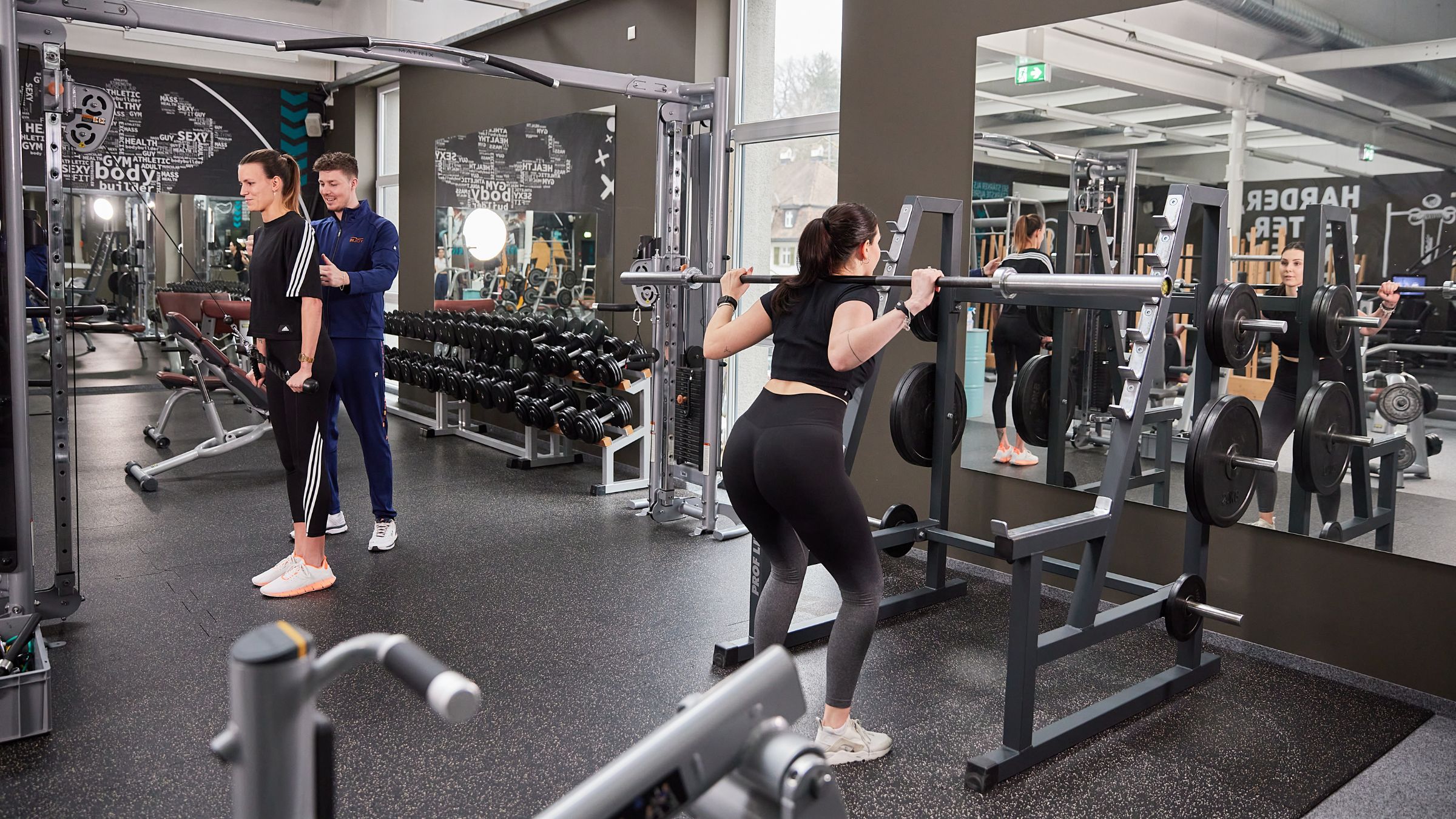 Eine Frau macht Kniebeugen mit einer Langhantel vor einem Spiel im INJOY Fitnessstudio Bregenz, während links daneben ein Trainer bei einer anderen Frau die Haltung korrigiert.