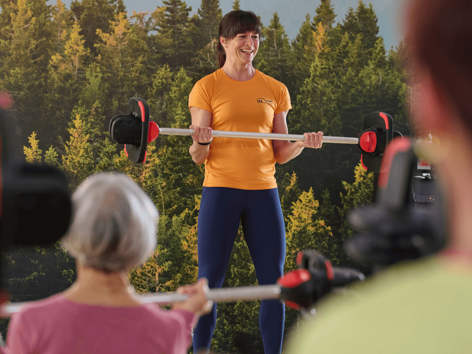 INJOY Kursleiterin in orangefarbenem Shirt leitet motiviert ein Langhantel-Workout an – Gruppenatmosphäre, Spaß und zielorientiertes Training im INJOY Kursbereich.