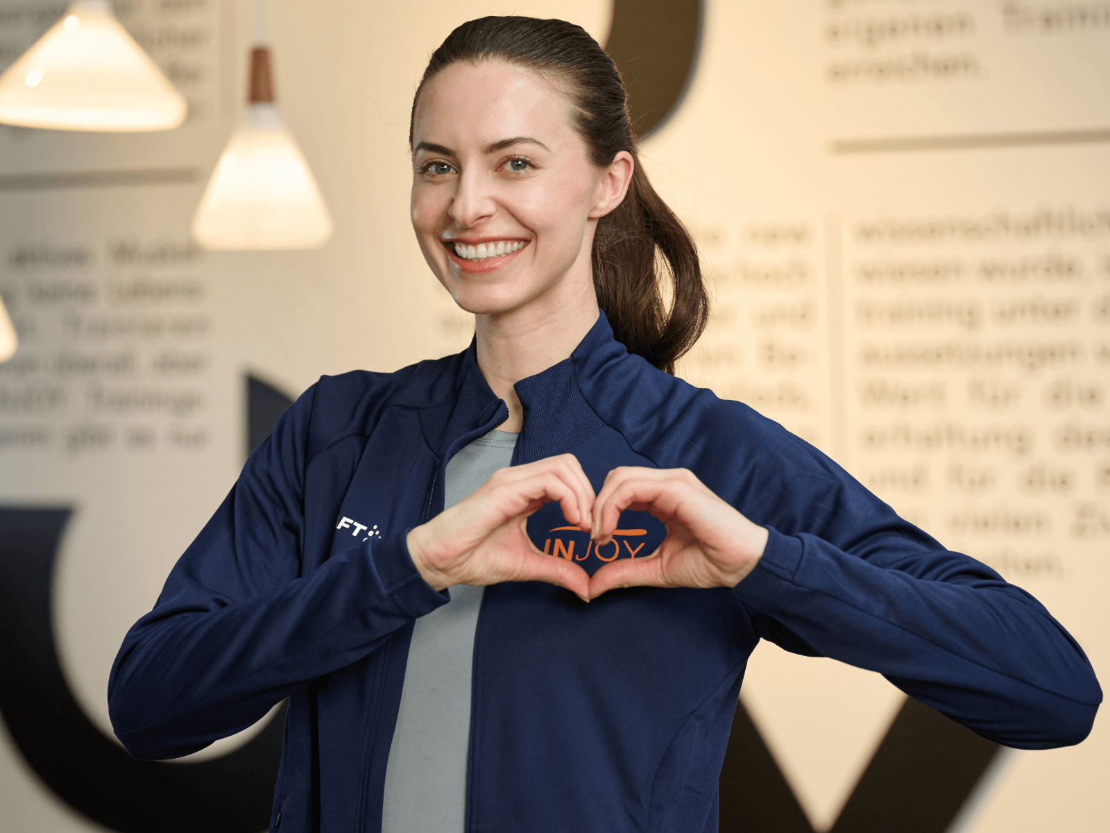 INJOY Trainerin formt ein Herz mit den Händen vor ihrem INJOY Logo auf der Jacke – Symbol für persönliche Zuwendung, Kompetenz und ganzheitliche Betreuung inklusive Ernährung.