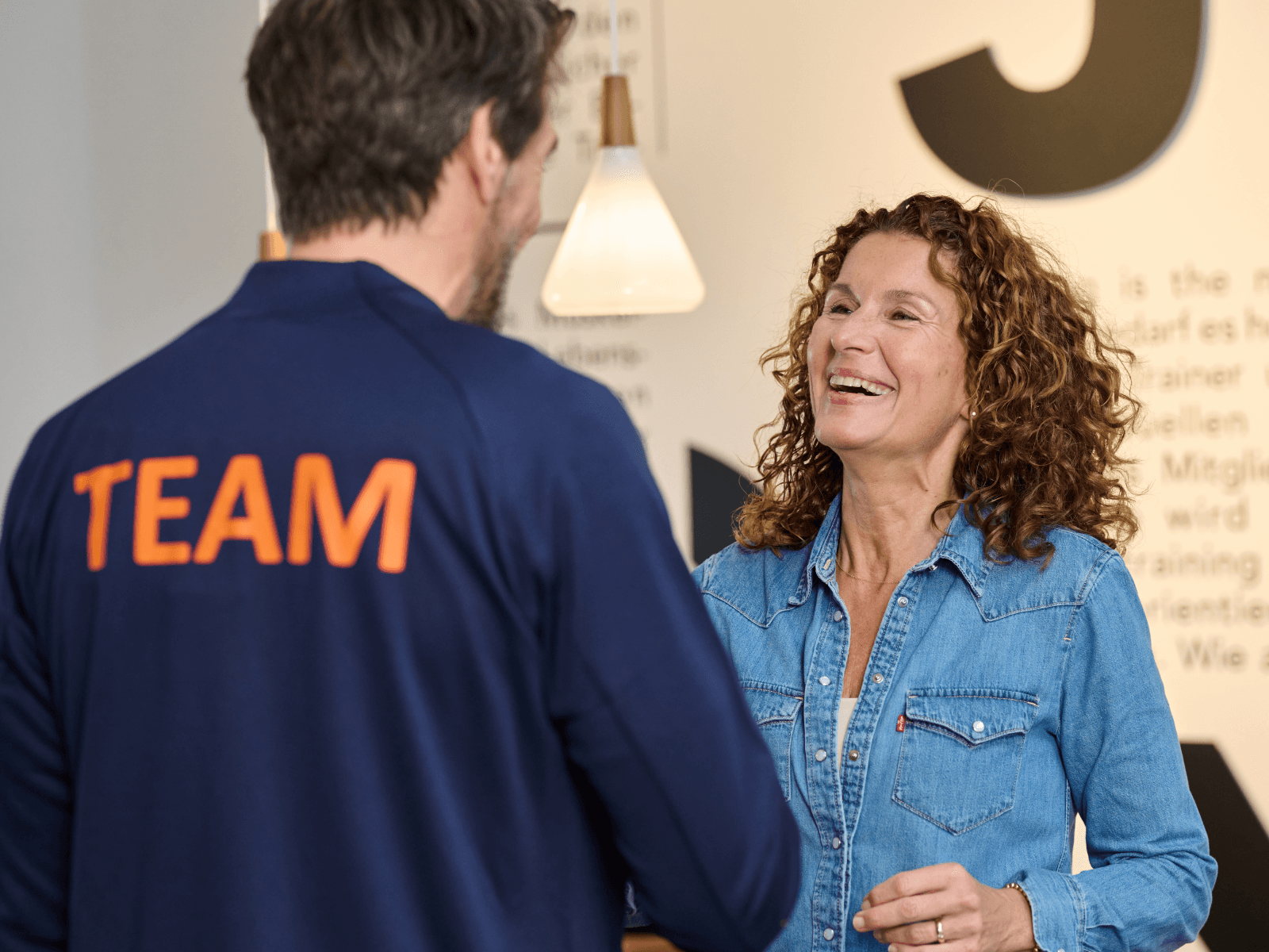 INJOY Trainer mit blauer TEAM-Jacke empfängt lachendes Mitglied zum ersten Termin der Ernährungsberatung