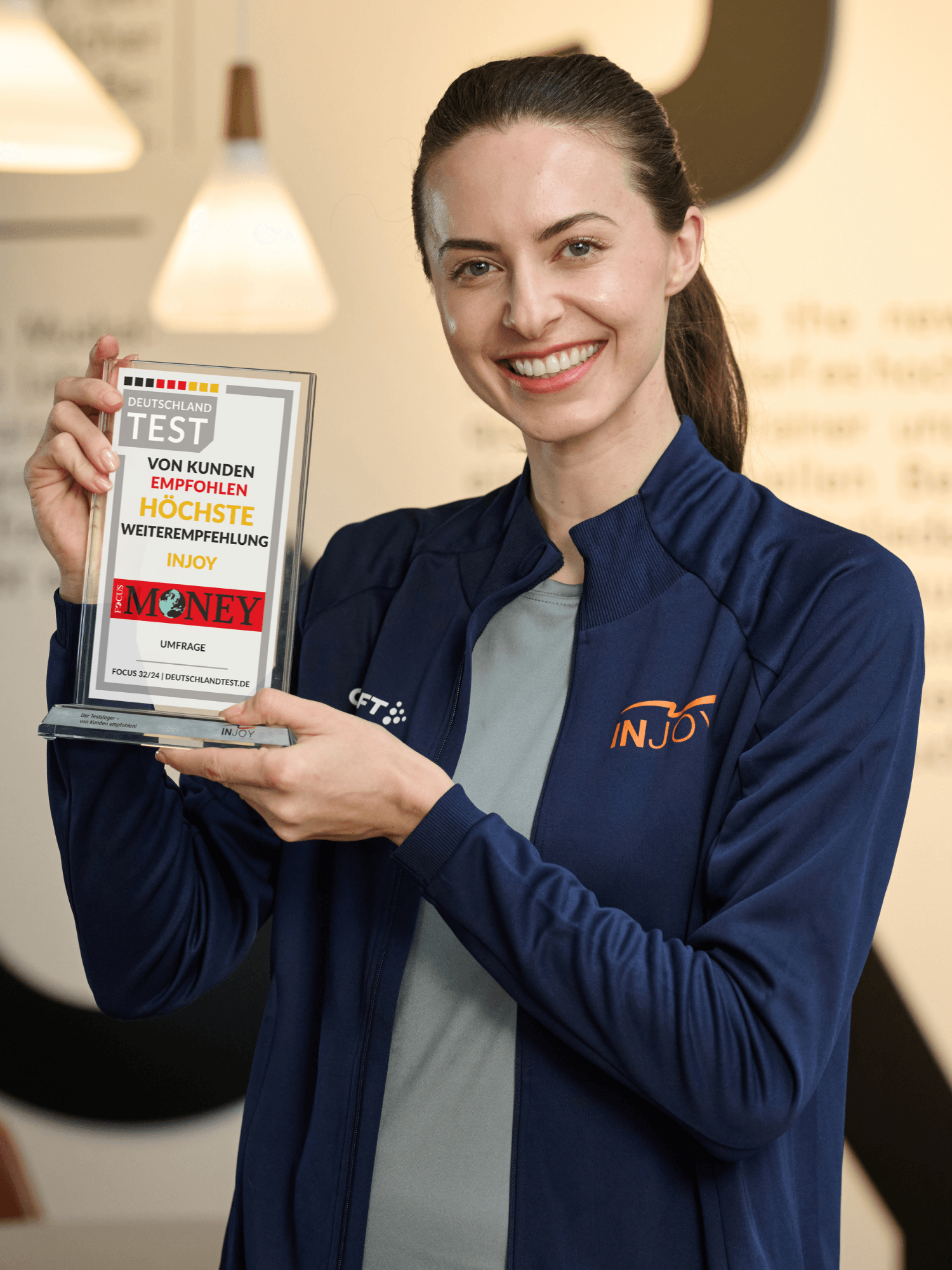 Sympathische INJOY Trainerin lächelt und hält stolz die Auszeichnung zum Weiterempfehlungs-Testsieger in die Höhe.