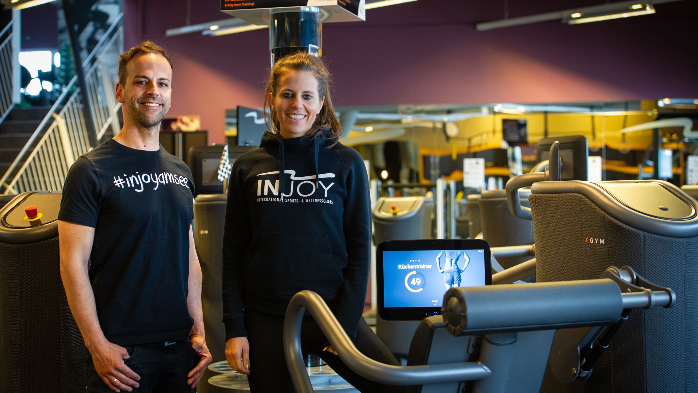 Ein Trainer und eine Trainerin auf einer Trainingsfläche im INJOY Fitnessstudio Saalfelden schauen lächelnd in die Kamera.