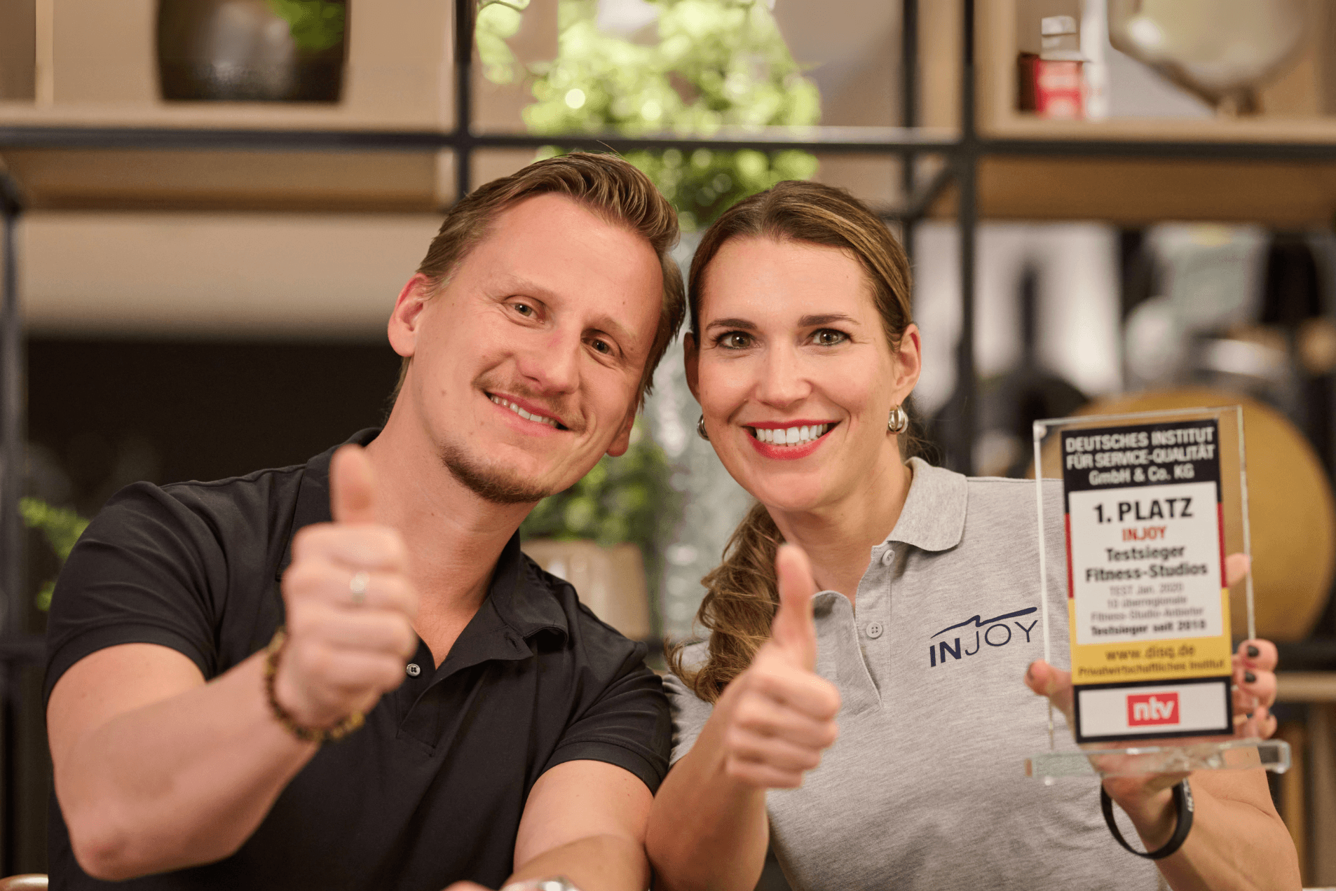 Eine INJOY Beraterin und ein INJOY Franchisenehmer zeigen beide einen Daumen hoch in die Kamera. Die Franchiseberaterin hält mit der anderen Hand den ntv Award für Platz 1 Testsieger Fitnessstudios hoch.
