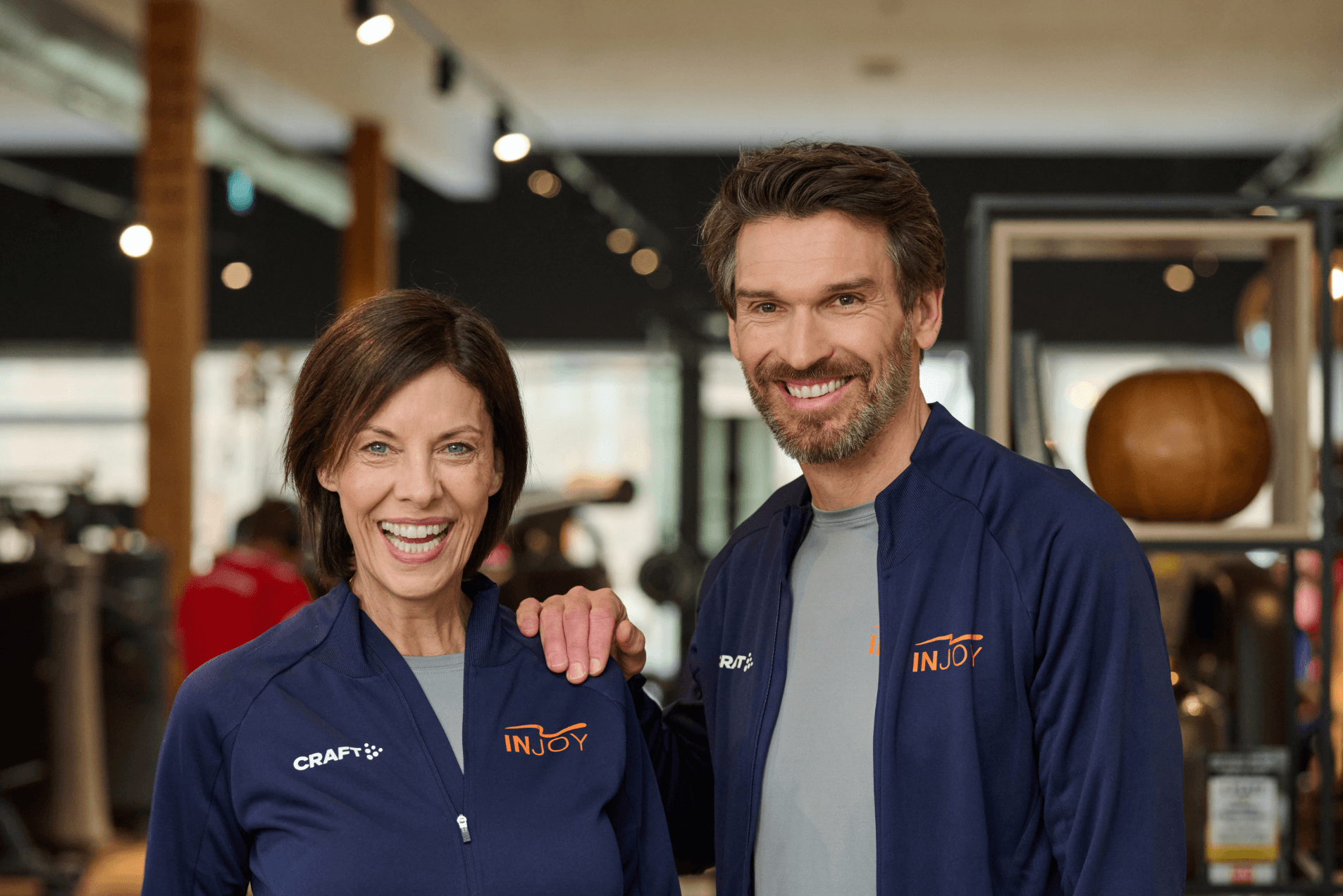 Eine INJOY Trainerin und ein INJOY Trainer stehen nebeneinander und lächeln in die Kamera.
