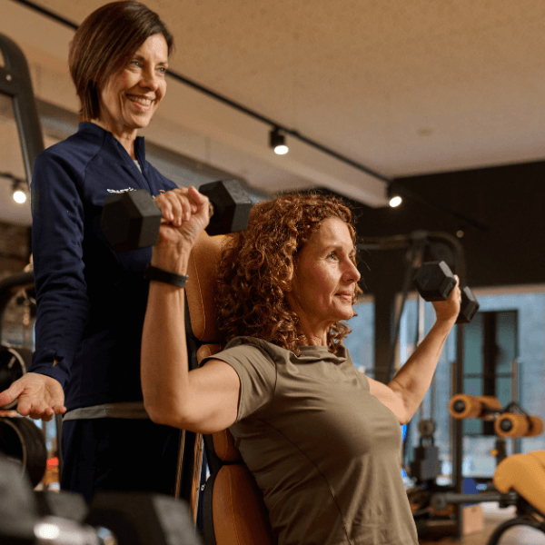 Weibliches INJOY Mitglied trainiert mit freien Gewichten ihre Schultern. Dahinter steht eine INJOY Trainerin und unterstützt.