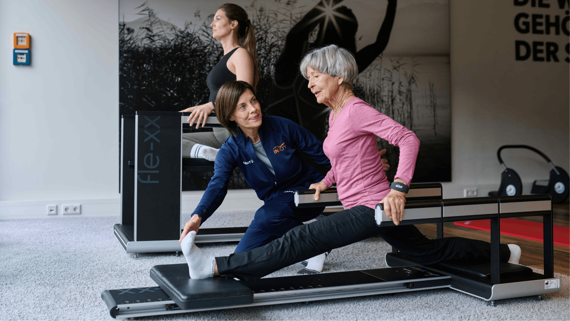 Ein älteres INJOY Mitglied führt eine Dehnübung durch und wird dabei von einer INJOY Trainerin betreut.