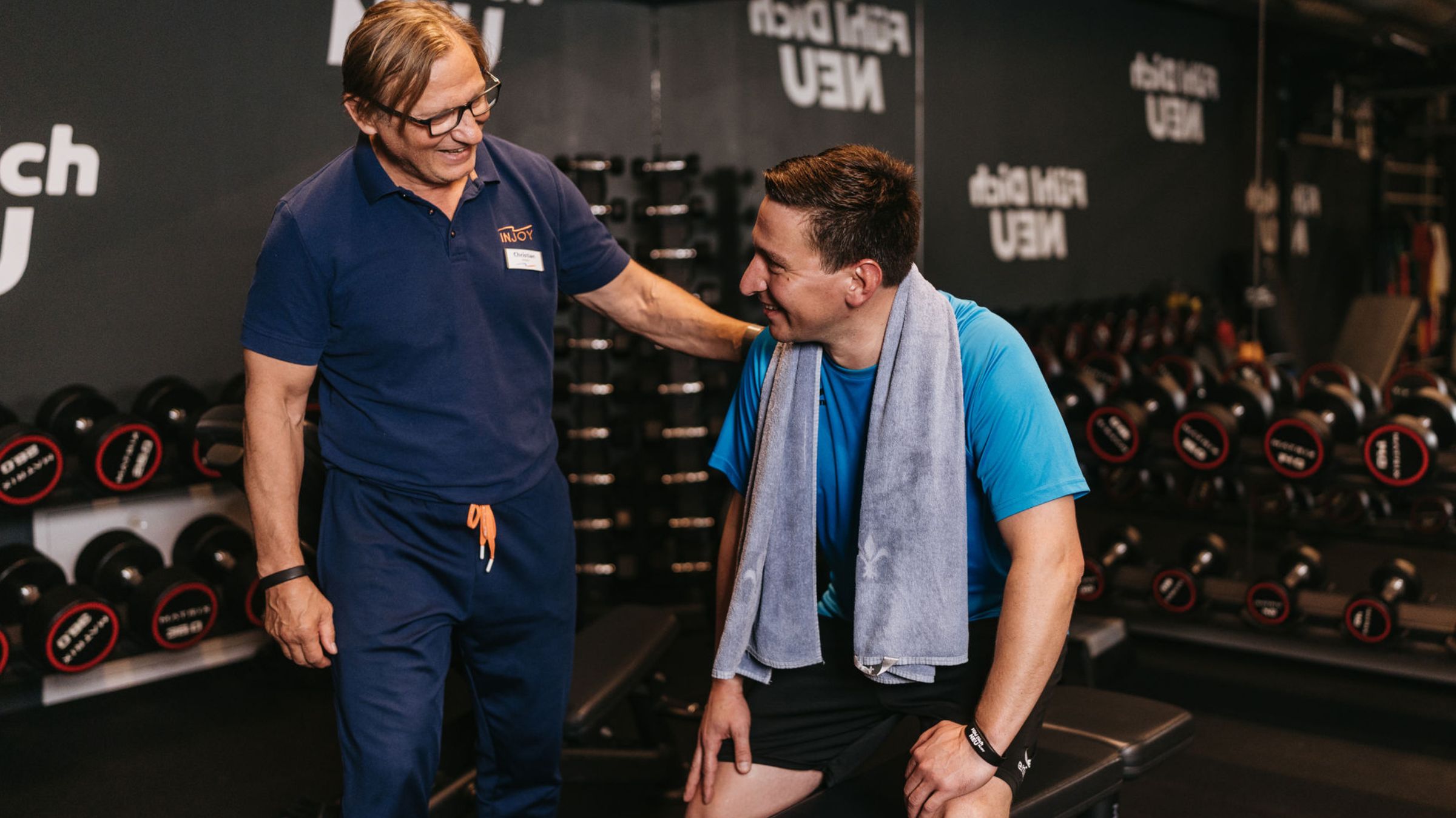 Ein Trainer legt seinen Arm lächelnd auf die Schulter eines Trainierenden im Freihantelbereich im INJOY Fitnessstudio Eferding.